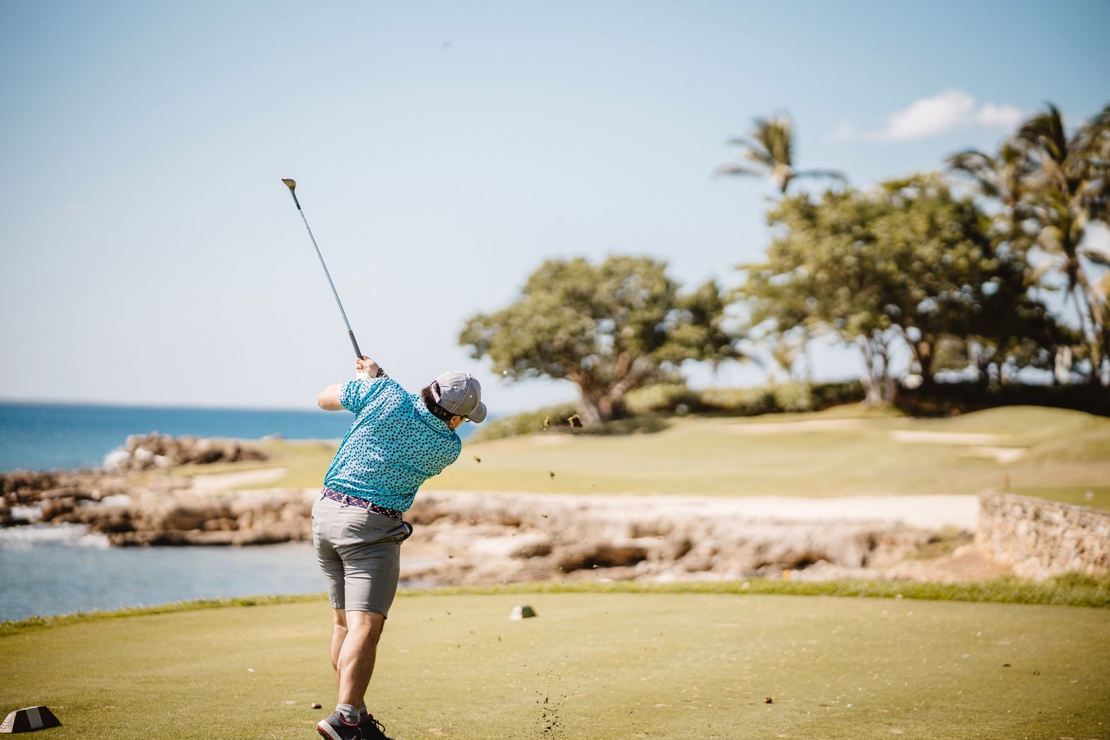 Golfer in a blue shirt teeing off beside a rocky shoreline with palm trees behind